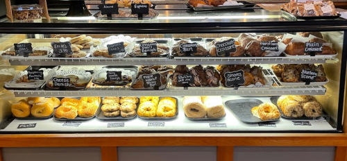 A display case filled with various baked goods, including pastries, scones, and bagels, labeled with chalkboard signs.