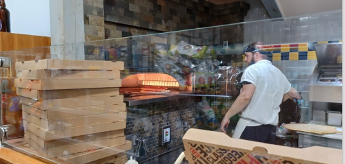 A chef prepares pizza in a restaurant kitchen, with a wood-fired oven and stacked pizza boxes in the foreground.