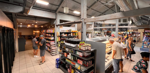 A busy market interior with shelves of products, shoppers browsing, and a café area in the background.