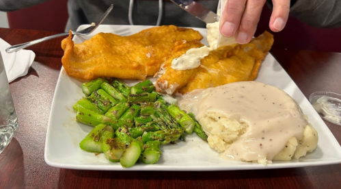 A plate with two fried fish fillets, mashed potatoes with gravy, and sautéed asparagus, with a hand adding sauce.