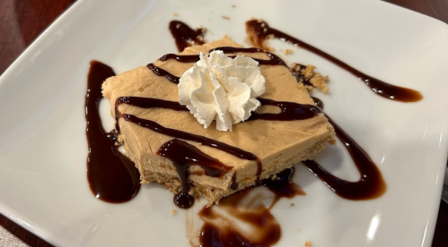 A slice of creamy dessert topped with whipped cream and drizzled with chocolate sauce on a white plate.