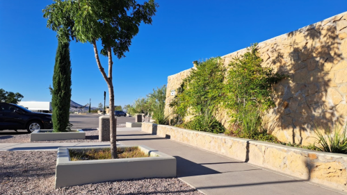 A sunny outdoor scene featuring a stone wall, trees, and a paved walkway in a landscaped area.