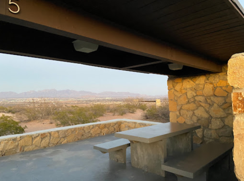 A stone picnic area with a table overlooks a desert landscape and distant mountains at sunset.