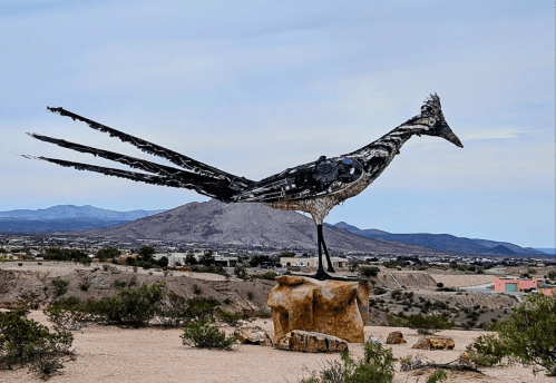 A large metal sculpture of a bird perched on a rock, set against a desert landscape with mountains in the background.