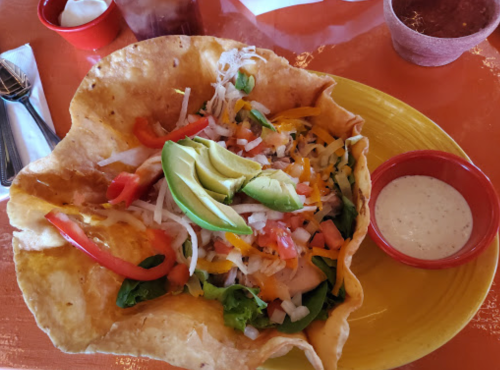 A colorful taco salad in a crispy shell, topped with avocado, tomatoes, and served with a side of dressing.