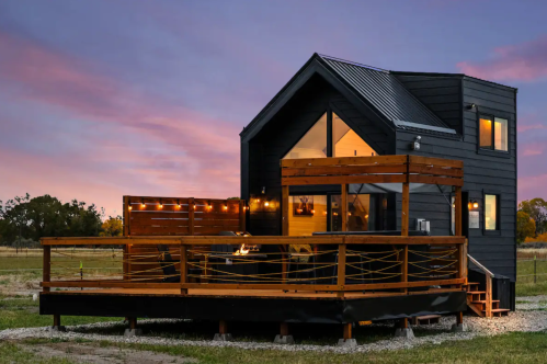 A modern black house with a wooden deck, surrounded by open fields under a colorful sunset sky.