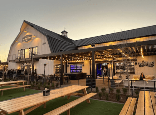 A modern barn-style restaurant with outdoor seating, string lights, and a cozy atmosphere at dusk.