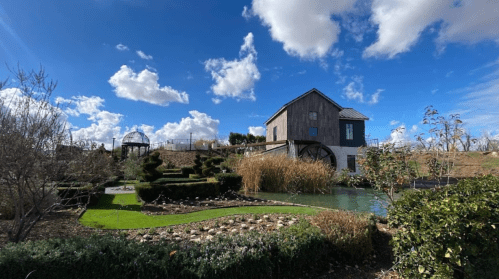 A scenic view of a house by a pond, surrounded by manicured gardens and blue skies with fluffy clouds.