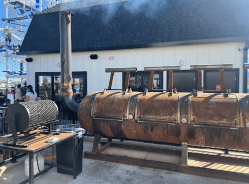A large, rusty smoker sits outside a building, with smoke rising from its chimney and people visible in the background.