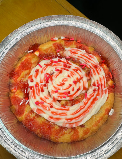 A circular dessert with a spiral of frosting and red sauce, topped with colorful sprinkles in a silver foil pan.