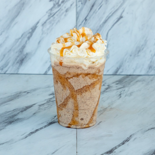 A creamy dessert drink topped with whipped cream and caramel drizzle, served in a clear cup on a marble surface.