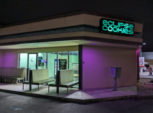 Exterior of Eclipse Cookies shop at night, featuring neon signage and seating outside.