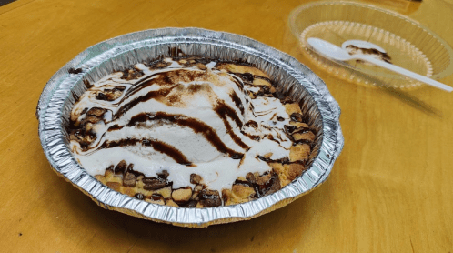 A dessert pie with a scoop of ice cream on top, drizzled with chocolate sauce, in a silver foil pie dish.