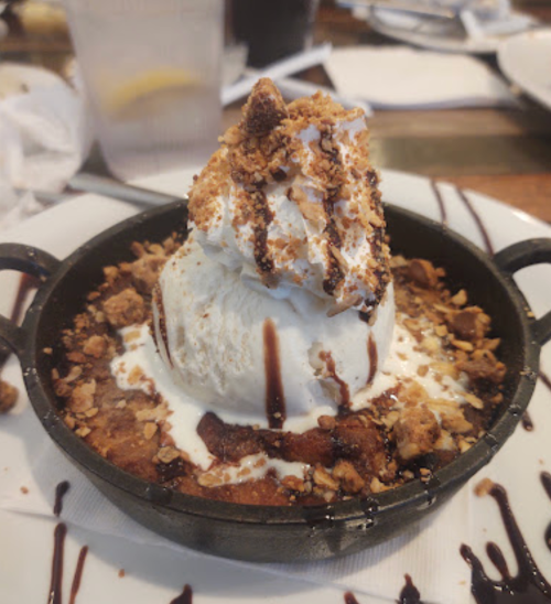A warm dessert in a skillet topped with ice cream, chocolate drizzle, and crushed nuts.