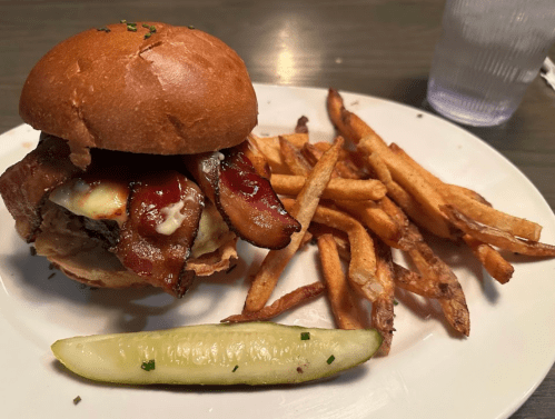 A juicy burger topped with bacon and cheese, served with crispy fries and a pickle on a white plate.