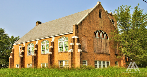 Abandoned brick church with boarded windows, surrounded by overgrown grass and trees under a clear blue sky.
