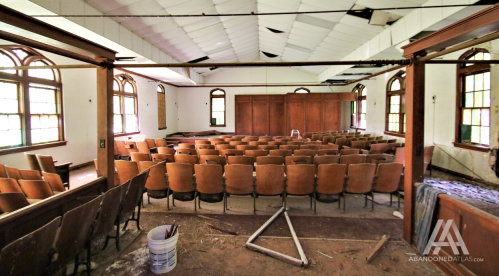 Abandoned auditorium with rows of empty wooden chairs and a dusty floor, featuring large windows and a wooden backdrop.