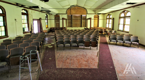 Abandoned interior of a hall with rows of empty chairs and deteriorating walls, illuminated by natural light.