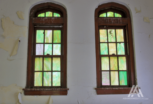 Two vintage windows with colorful stained glass, framed by peeling paint on the walls. Natural light filters through.