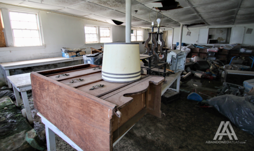 An abandoned room filled with clutter, featuring an old wooden table and a lamp on top. Dust and debris are scattered around.