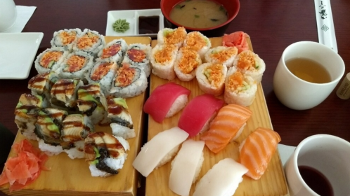 A wooden platter with various sushi rolls, sashimi, pickled ginger, miso soup, and a cup of tea.