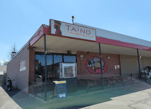 Exterior of Taíno Smokehouse, featuring a red sign and mural, with outdoor seating and a clear blue sky.