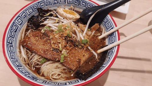 A bowl of ramen with sliced pork, soft-boiled egg, bean sprouts, and green onions, served with chopsticks.