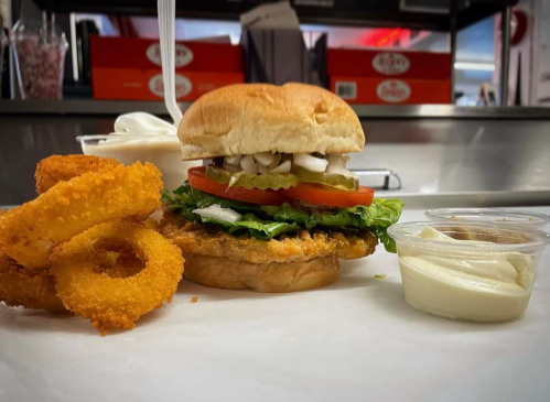 A crispy chicken sandwich with lettuce, tomato, pickles, and onion, served with onion rings and dipping sauce.