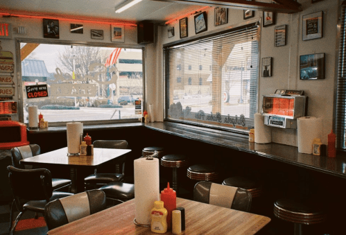 Retro diner interior with tables, condiments, and vintage decor, featuring large windows and a jukebox.