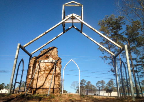 A skeletal metal structure frames the ruins of a stone building against a clear blue sky and trees in the background.