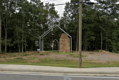 A partially constructed building frame stands among trees, with a stone wall remaining, beside a road.
