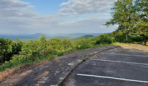 A scenic view of rolling hills and mountains under a partly cloudy sky, with an empty parking area in the foreground.