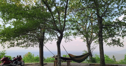 A hammock hangs between two trees, with a motorcycle nearby and a scenic view of mountains in the background.