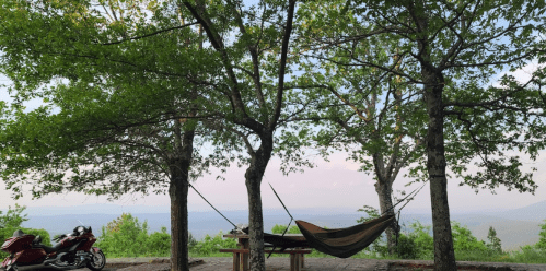 A hammock hangs between two trees, with a scenic view of mountains in the background and a motorcycle nearby.