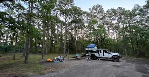 A campsite with a Jeep, a campfire, and a person sitting by a picnic table surrounded by trees at dusk.