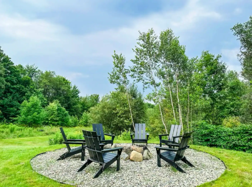 A circular seating area with black chairs around a fire pit, surrounded by greenery and trees under a cloudy sky.