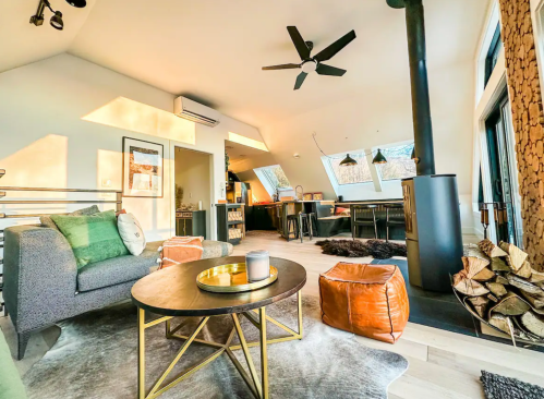 Cozy modern living space with a sofa, coffee table, and wood stove, featuring large windows and warm lighting.