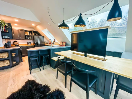 Modern kitchen with black cabinetry, a wooden dining table, and a TV on a stand, featuring large windows for natural light.