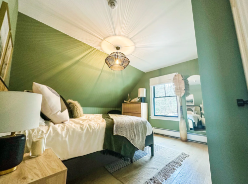 Cozy bedroom with green walls, a bed, decorative mirror, and warm lighting from a stylish ceiling fixture.