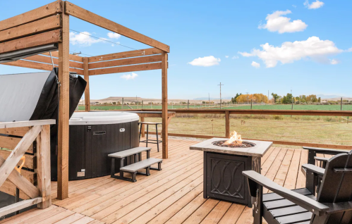 A wooden deck with a hot tub, fire pit, and seating, overlooking a grassy field and blue sky.