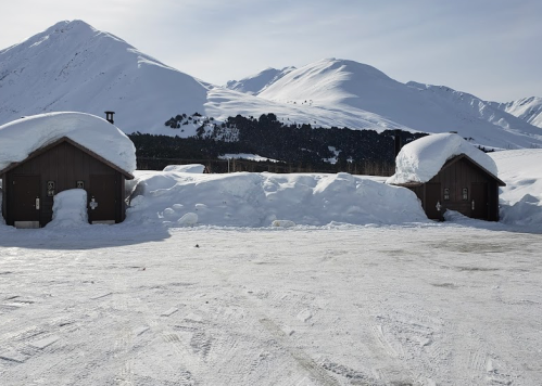 Two wooden cabins covered in snow, surrounded by a snowy landscape and mountains under a clear sky.