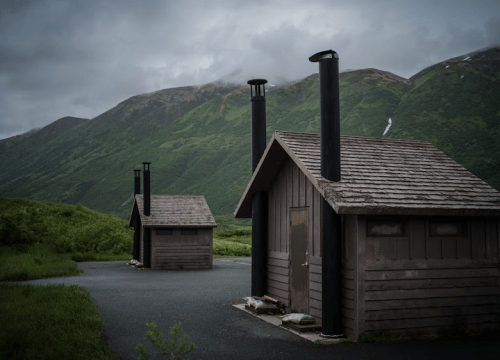 Two rustic wooden cabins with black chimneys, set against a backdrop of green mountains under a cloudy sky.