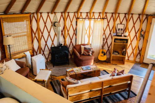 Cozy yurt interior with a seating area, guitar, wood stove, and natural light from large windows.