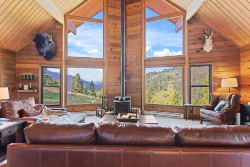 Cozy wooden living room with large windows, mountain views, and rustic decor, including mounted animal heads.