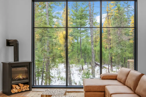 Cozy living room with a large window showcasing a forest view of trees and snow, featuring a fireplace and sofa.