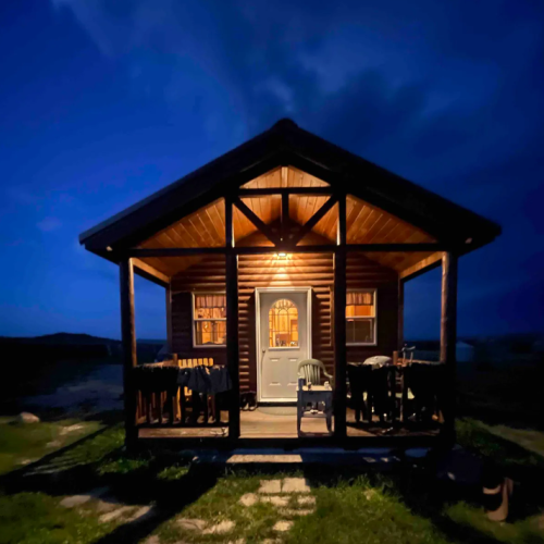 A cozy wooden cabin illuminated at night, with a welcoming front porch and a warm glow from the windows.