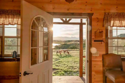 A wooden door opens to a scenic view of a field and mountains at sunset, with sunlight streaming in.