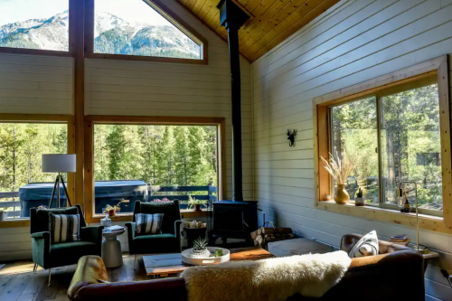 Cozy cabin interior with large windows, mountain views, and a wood stove, featuring comfortable seating and natural decor.