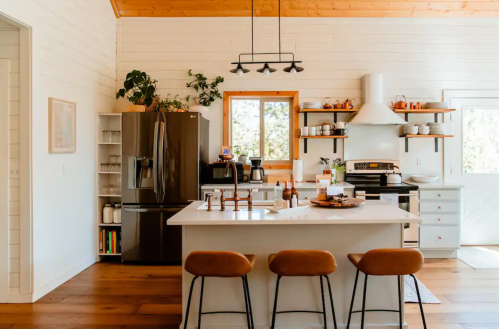 Modern kitchen with white walls, wooden accents, a large island, and stylish bar stools, featuring plants and open shelving.