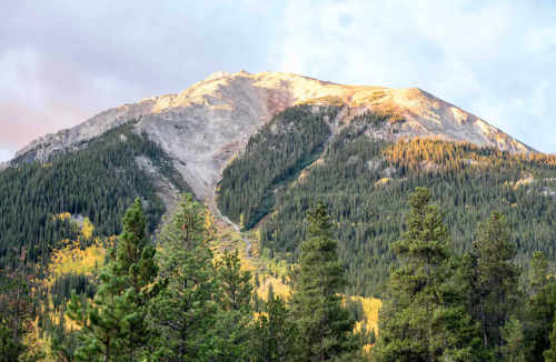 A mountain peak bathed in sunlight, surrounded by lush green trees and a clear sky.